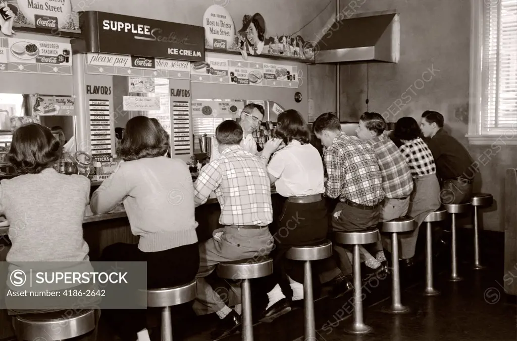 1950S Rear View Of Group Of Teenage Boys & Girls Sitting Together At A Soda Fountain Malt Shop Counter Snack Food