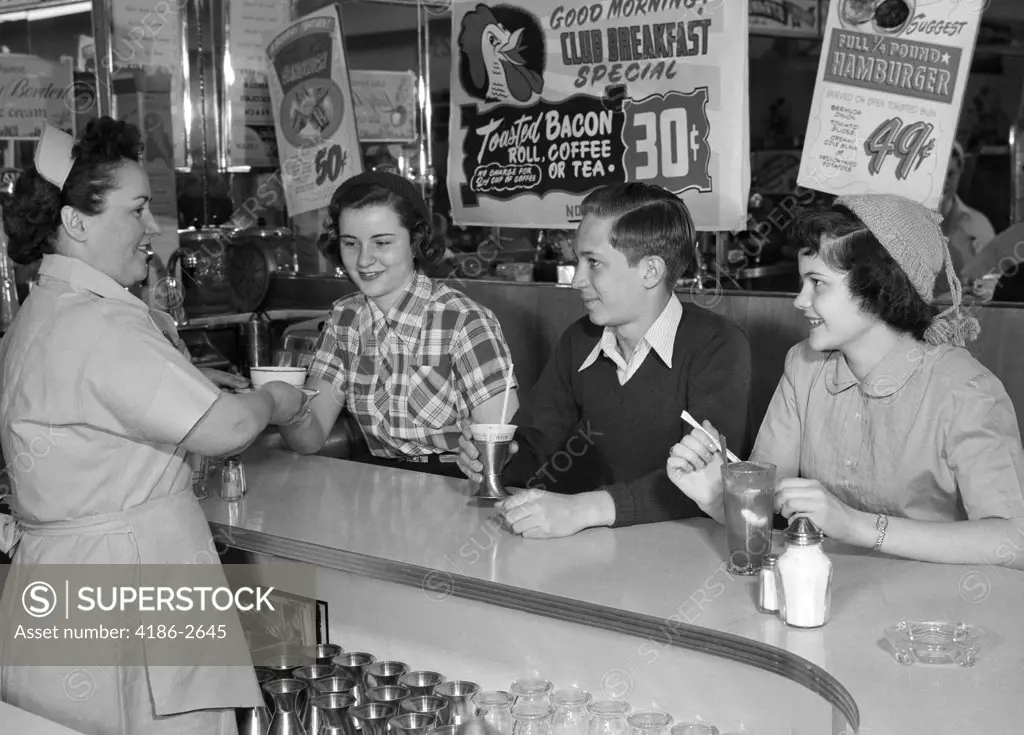 1950S Smiling Woman Waitress Serving Two Teenage Girls And One Boy At Diner Counter