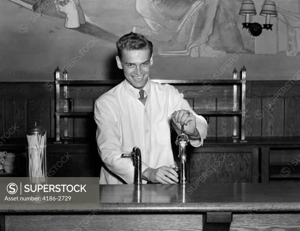 1940S Soda Jerk Smiling While Filling A Glass From The Fountain Behind The Counter Dressed In Shirt Tie And White Smock