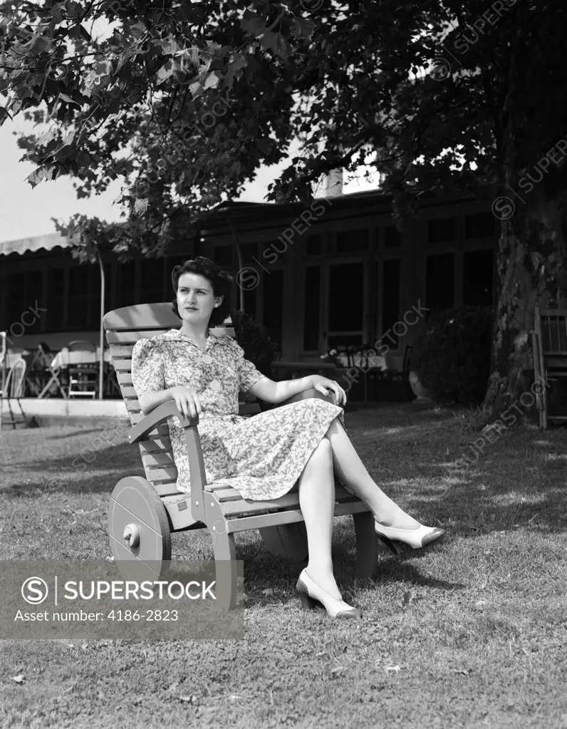 1940S Woman Sitting In Wooden Lawn Chair With Wheels On Lawn In Front Of House Looking Off Into Distance