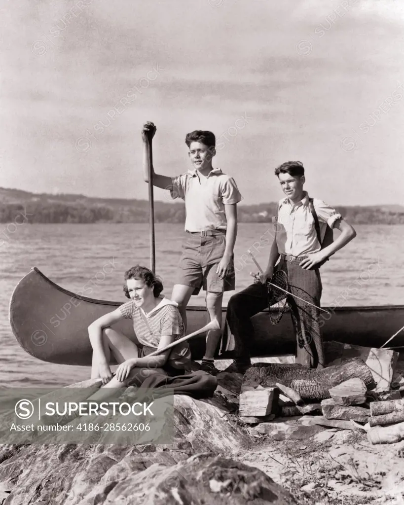 1930s GROUP OF TEENS GIRL & TWO BOYS WITH CANOE AND FISHING GEAR SURVEYING  THE ROUTE AHEAD SITTING ON ROCKS BY LAKE - SuperStock