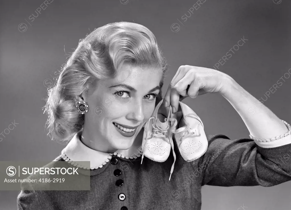 1950S 1960S Blond Woman Communicating That She Is Mother To Be By Holding Up A Pair Of Baby Shoes And Smiling Studio Symbolic