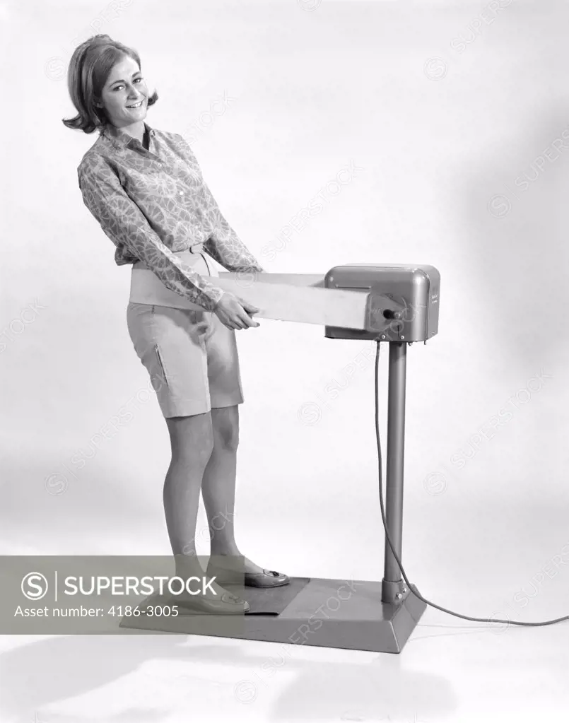 1960S Smiling Young Woman Standing On Weight Reducing Vibrating Exercise Machine Looking At Camera