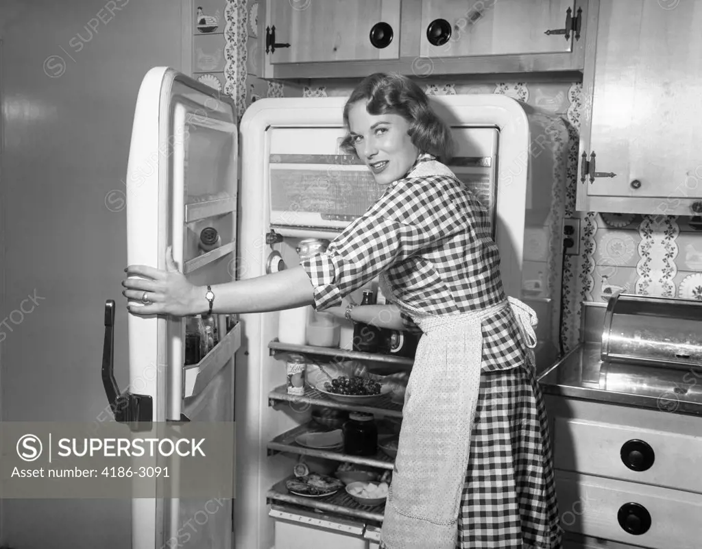 1950S Woman Wearing Gingham Dress And Apron Opening Refrigerator Door