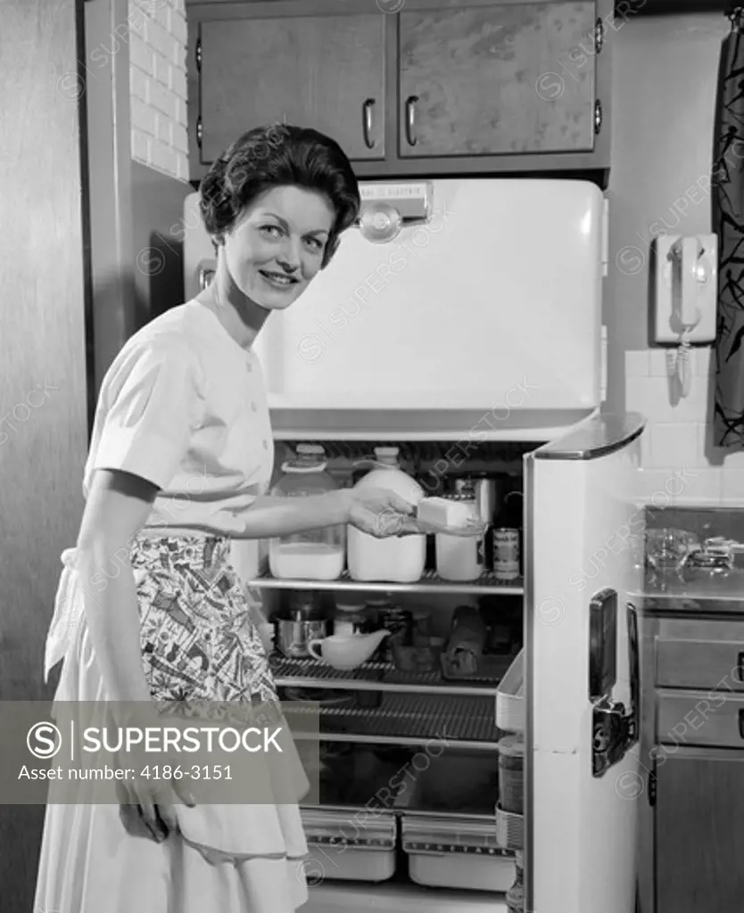 1950S Smiling Woman Housewife Putting Stick Of Butter Into Electric Refrigerator In Kitchen Looking At Camera