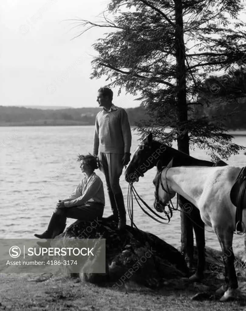 1930S Man Standing On Rock Lakeside Holding Reins Of Two Horses With Woman Seated Beside Him Looking Off To Side