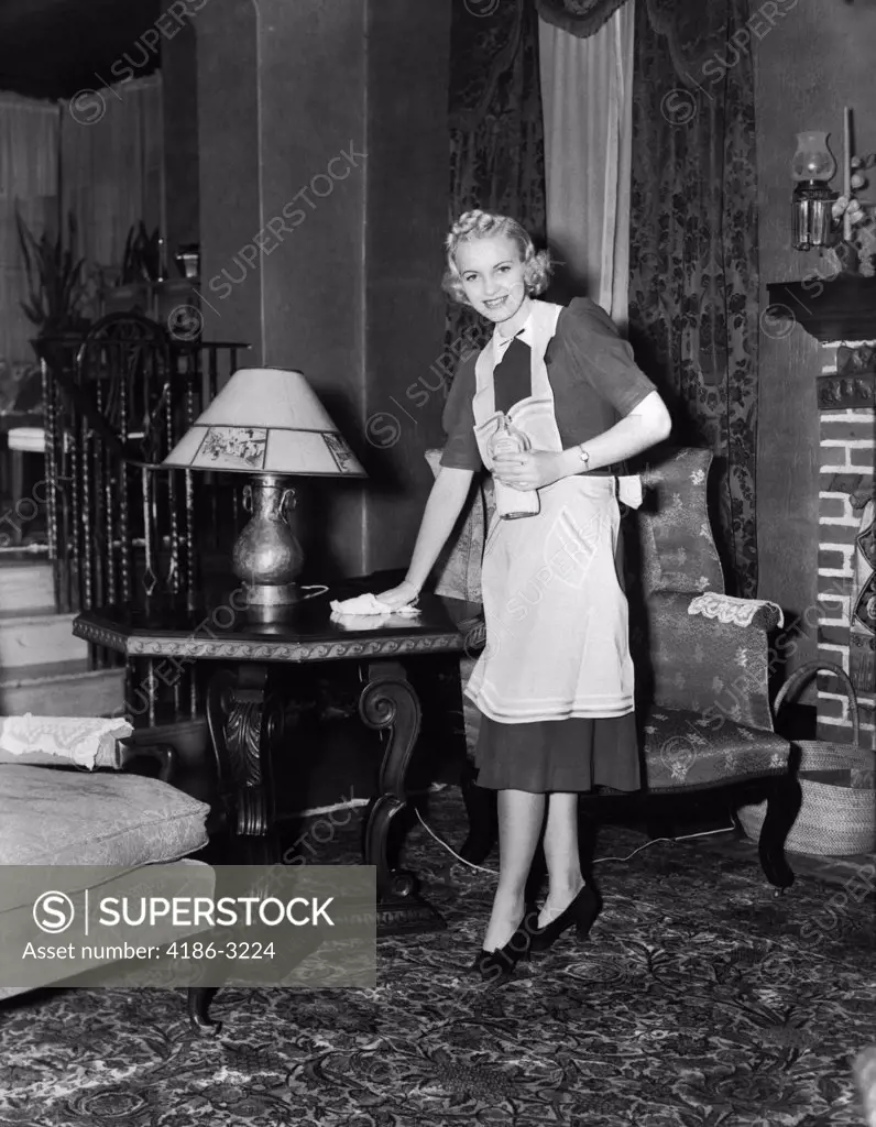 1940S Blonde Woman Housewife Maid Wearing Apron Cleaning Polishing Wooden End Table In Ornate Living Room