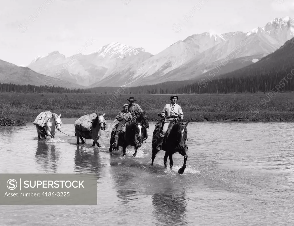 1930S Two Women Man Riding Horseback Through Shallow Water Two Extra Horses Carry Camping Equipment Mountains In Background