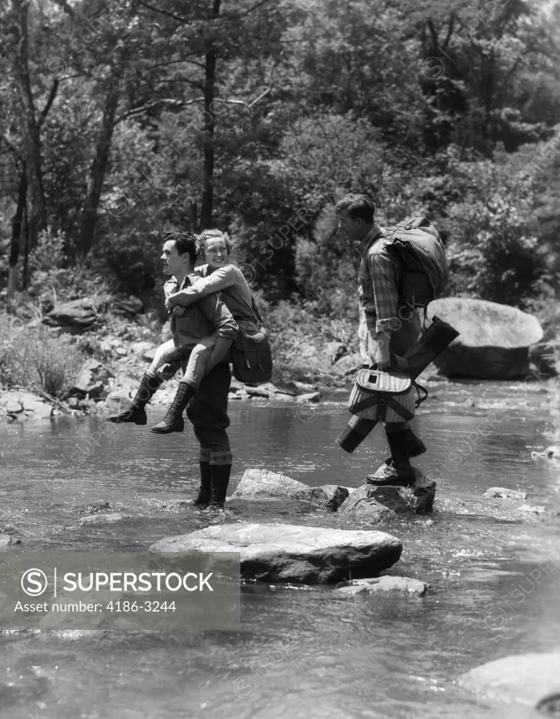 1930S 3 People Crossing Fording Stream One Man Carries Woman Piggy Back Other Carries Angling Fishing Gear Stepping Stones Water