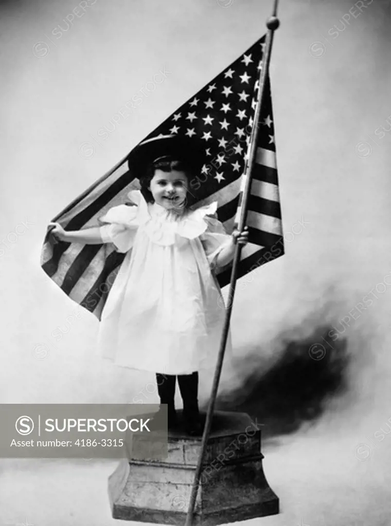 1890S Turn Of Century Smiling Little Girl Standing On Platform Wearing A White Dress And A Dark Hat Holding A American Flag