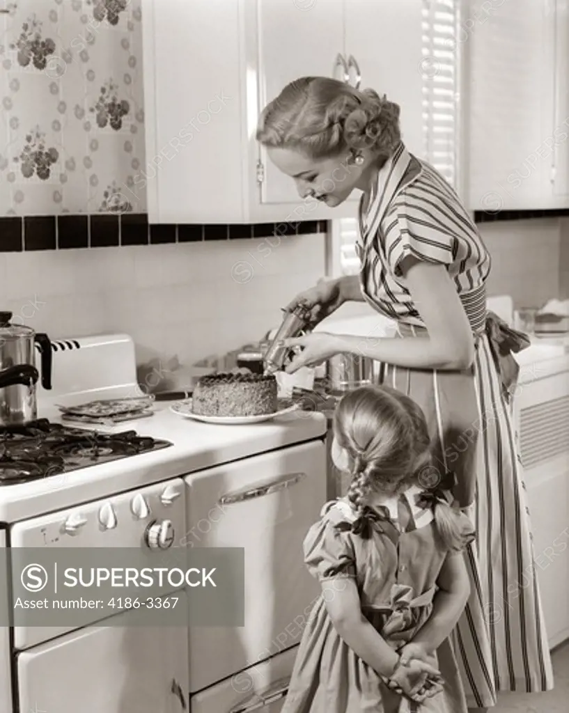 1950S Housewife In Kitchen Decorating Cake On Stove With Pastry Gun While Little Daughter Watches
