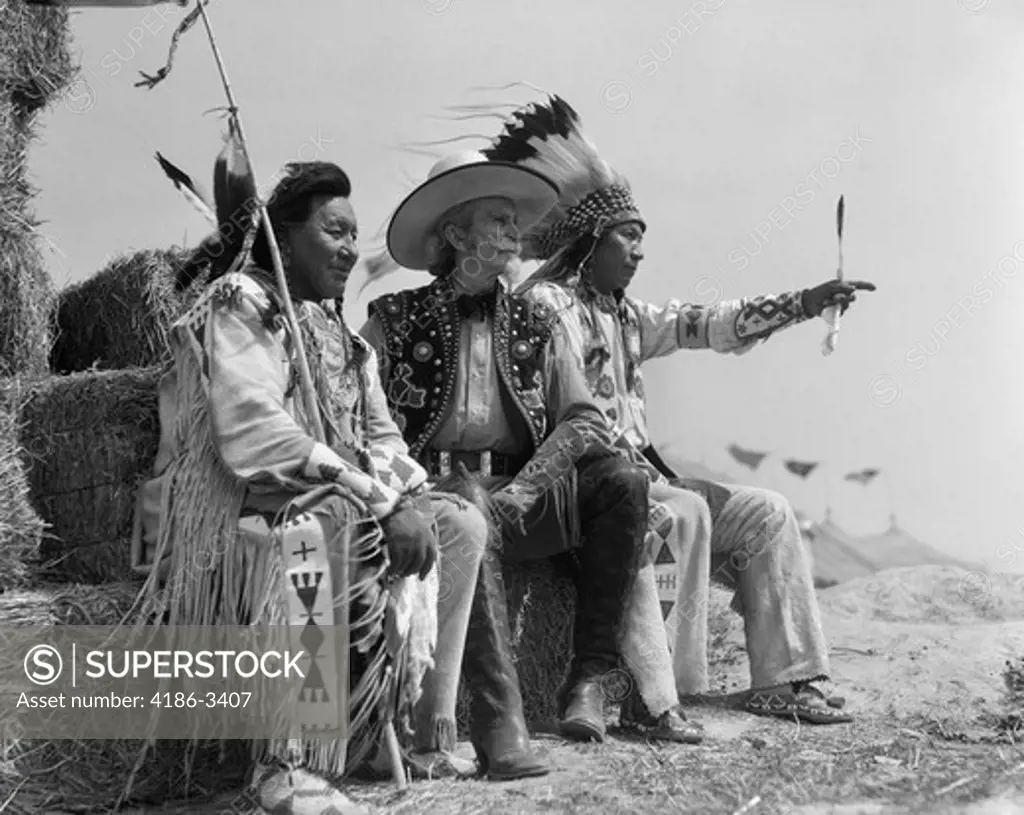 1940S Pair Of Indians In Full Costume Sitting On Bales Of Hay With Cowboy Between Them