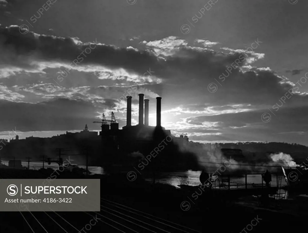 1930S Sunrise Sunset Behind Silhouette Of Industrial Power Plant With Smoke Stacks Near River And Railroad Tracks Outdoor