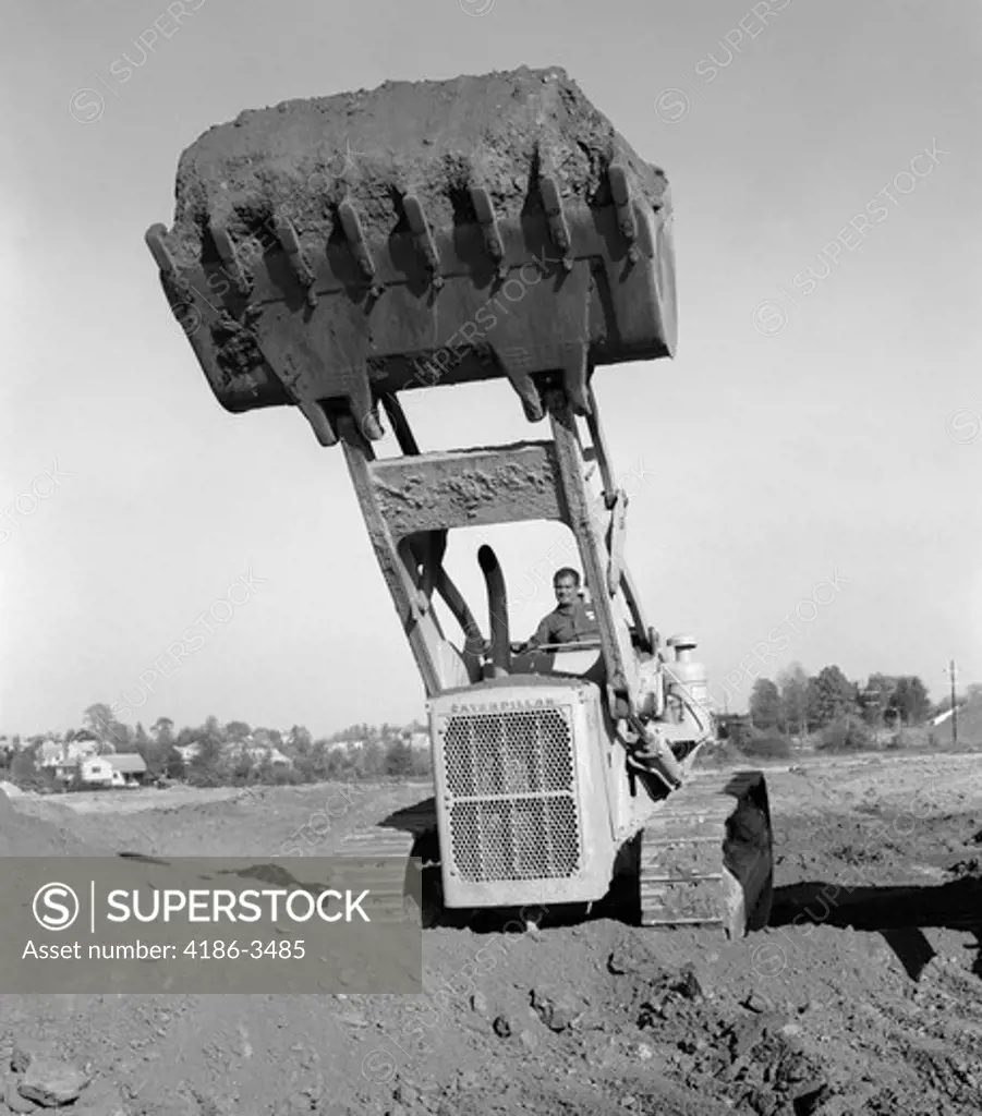 1960S 1970S Man Operating Caterpillar Heavy Machinery Bull Dozer Lifting Shovel Full Of Dirt Soil Construction Site