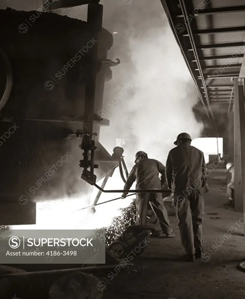 1960S Pair Of Steel Workers In Construction Hats Stirring & Stoking Molten Material Pouring From Large Container