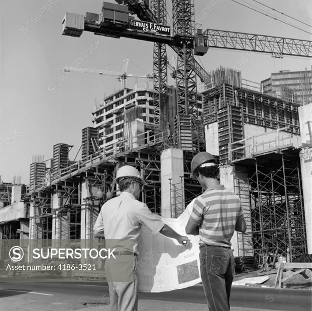 1980S Rear View Of Two Hard Hat Workers Standing In Front Of Major Construction Site Looking Over Building Plans Men Outdoor