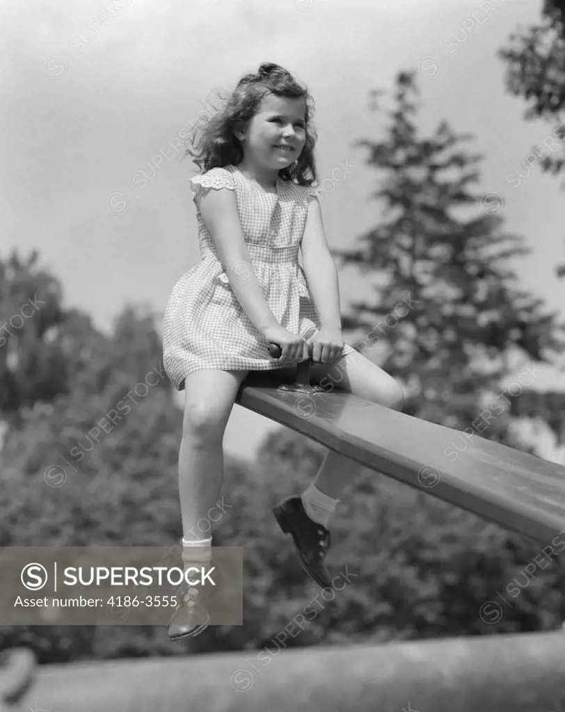 1950S 7 Year Old Girl Wearing Dress Sit On Seesaw Teeter Totter In Up Position In Park Smiling Fun Playground Play Up And Down