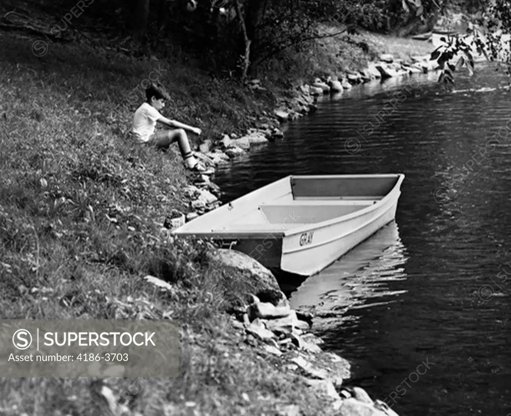 1960S Side View Of Boy Sitting On Bank Of Lake Next To Rowboat In Water