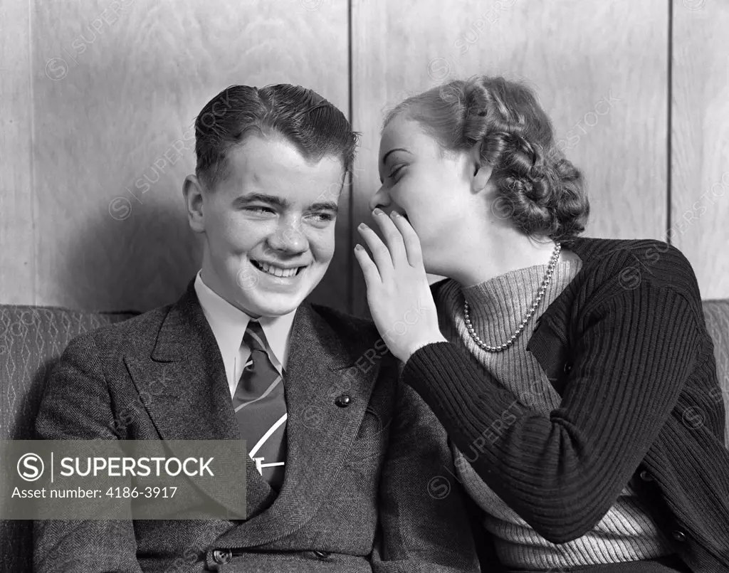 1930S 1940S Young Teenage Couple Sitting On Couch Girl Whispering Secret Into Smiling Boy'S Ear