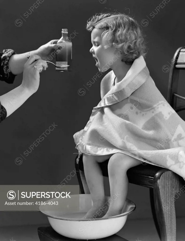 1940S Sick Little Girl Sitting In Chair Wrapped In Blanket Feet Soaking In Hot Water Taking Medicine On Spoon From Mother