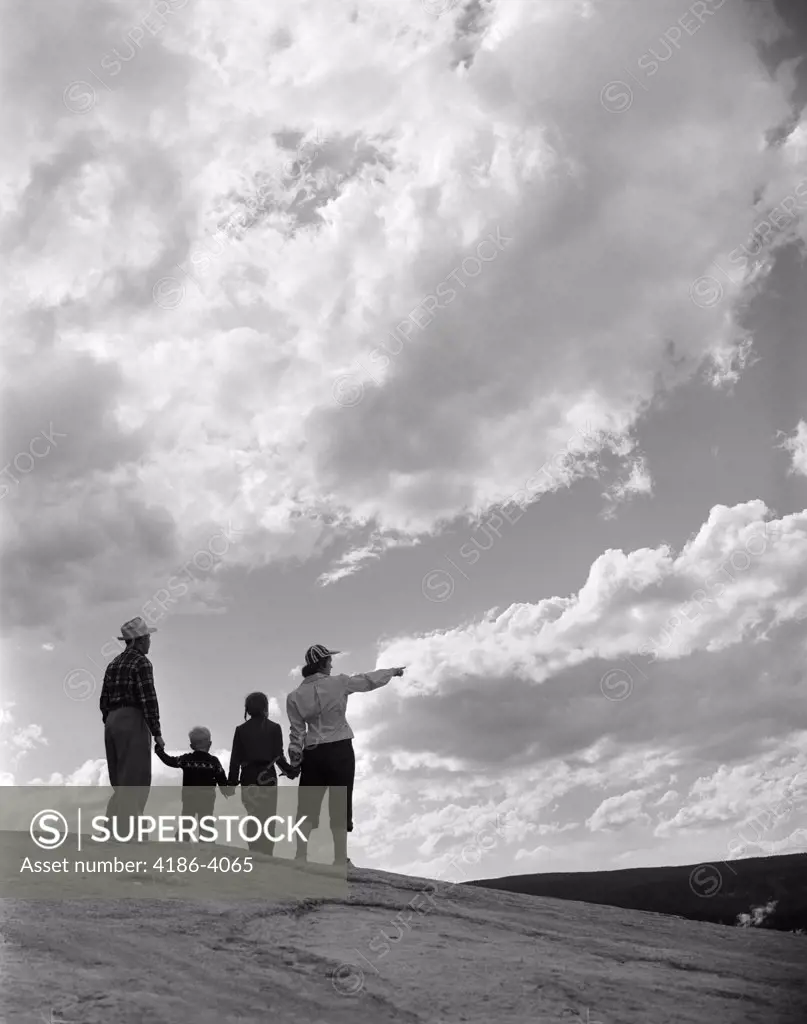 1950S 1960S Family Of Four Silhouetted On Top Rocky Hill Pointing At Clouds In Sky