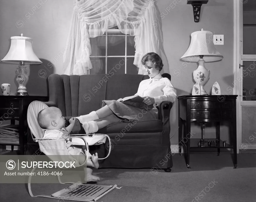 1950S Teen Babysitter Seated On Sofa Reading Schoolbook While Baby In Bouncy Sling Chair Grabs Her Propped Up Foot