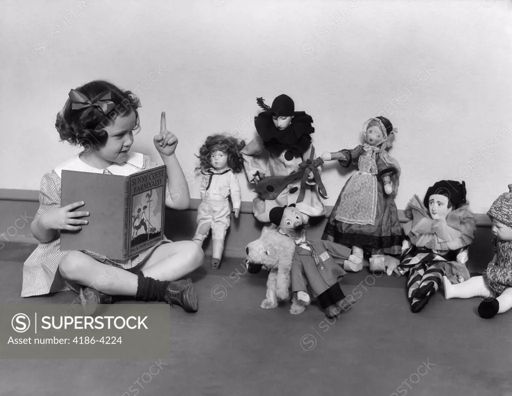 1930S Little Girl Pointing A Finger Reading Book To A Row Of Dolls Toys Stuffed Animals