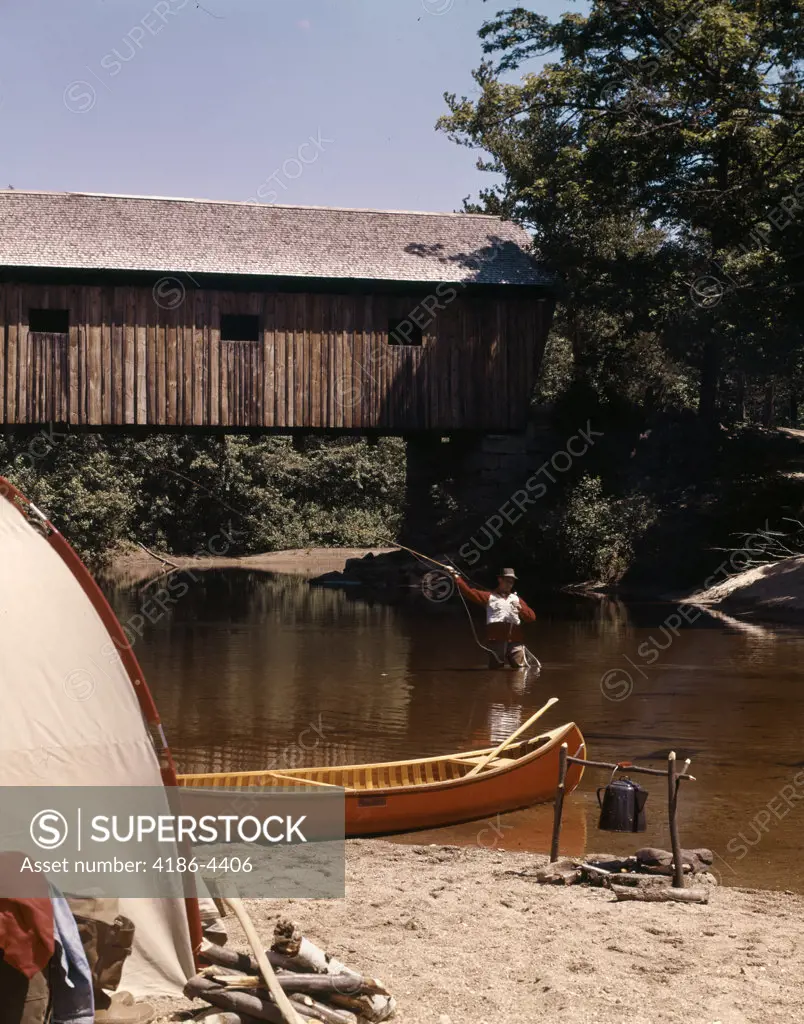 1960S 1970S Man Fly-Fishing Standing In Stream By Covered Bridge And Campsite With Tent Canoe Campfire