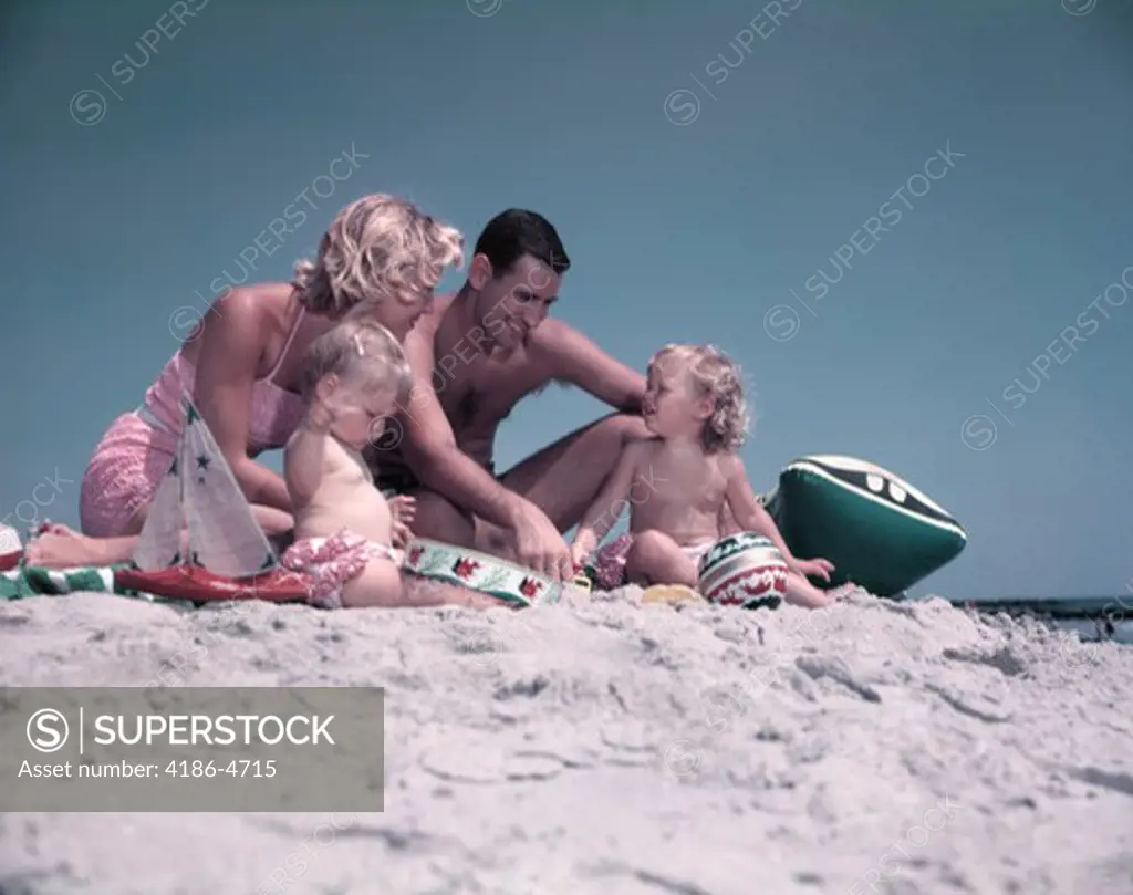 1950S Family Sitting On Sandy Beach Man Woman Mother Father Two Children Boy Girl Playing Sand Toys Summer Vacation