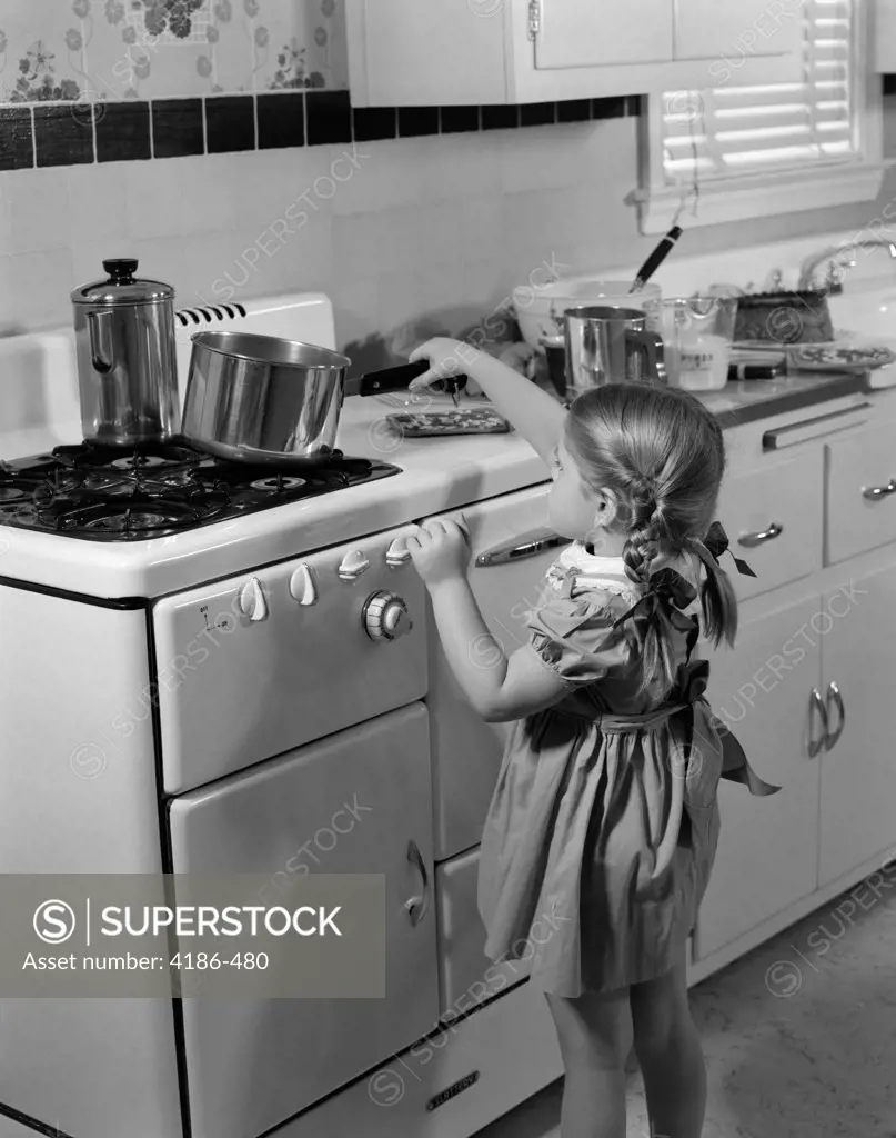 1950S Girl Toddler Taking Hot Saucepan Off Of Stove Just Below Her Eye Level