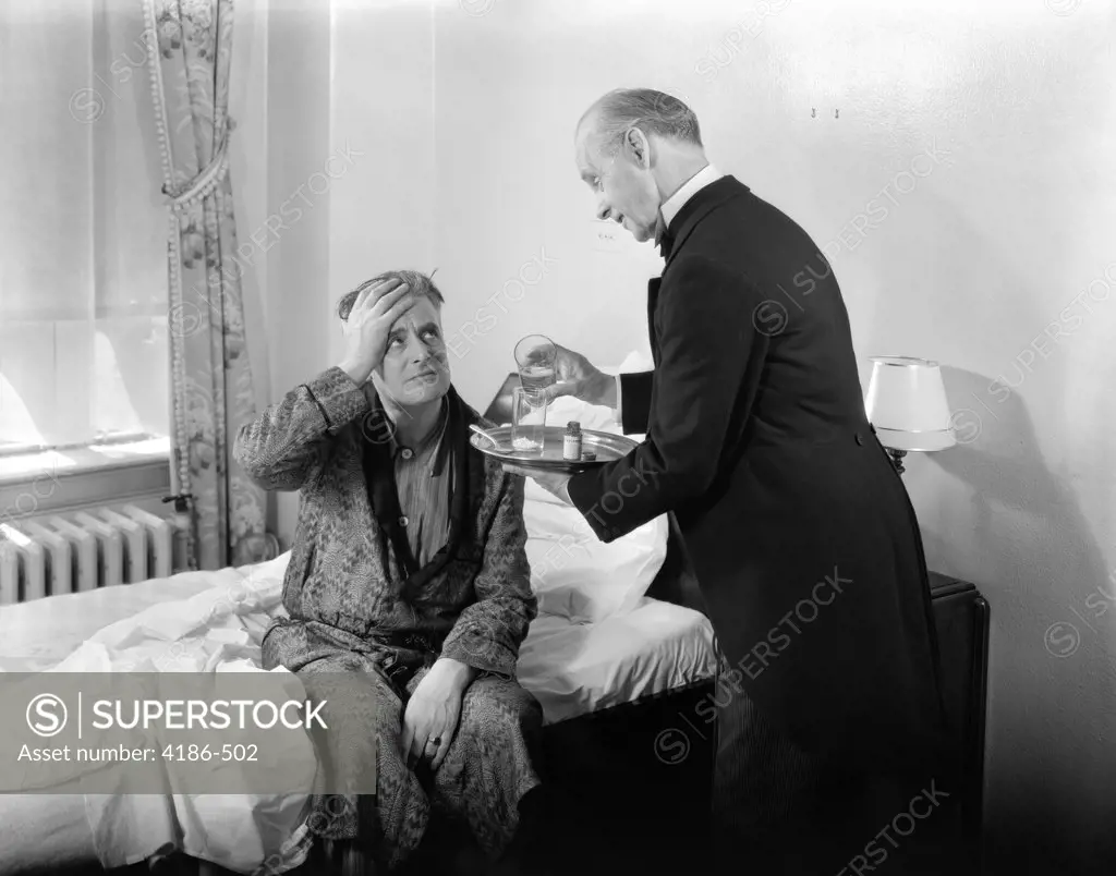 1930S 1940S Man Sit On Bed Hand To Head Doctor Butler With Tray Mixing Powder In Glasses Headache Hangover Pain