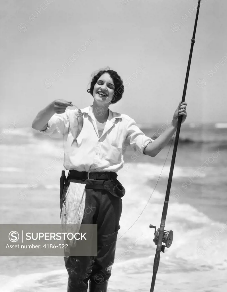 1920S 1930S Smiling Woman Standing In Ocean Surf Wearing Rubber Waders Holding Fish And Fly Fishing Rod