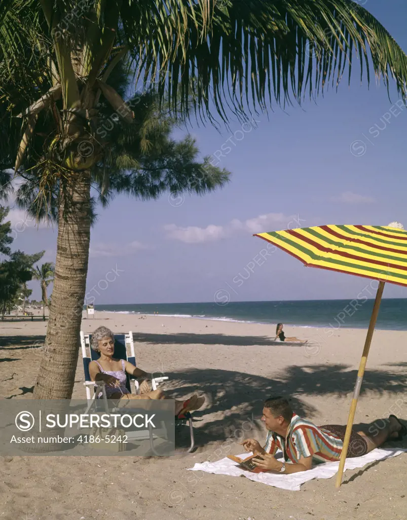 1960S Man And Woman Couple Sunbathing On Beach In Florida Talking Together Under Palm Tree And Colorful Beach Umbrella