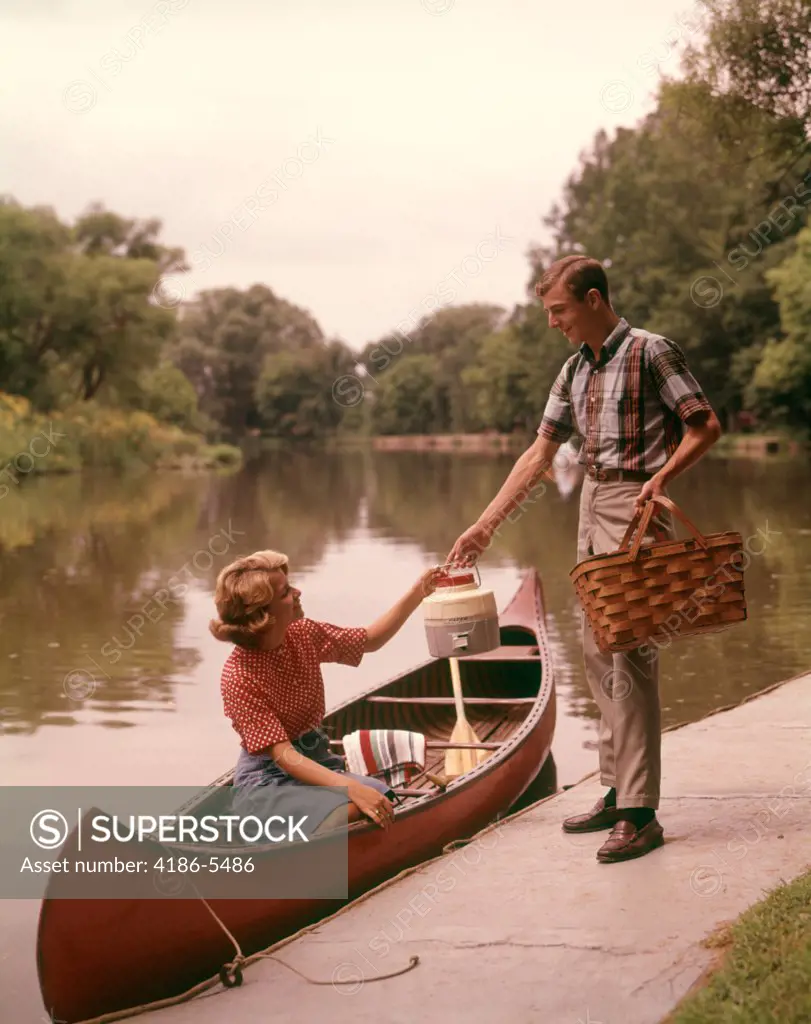 1960S Young Couple Loading Picnic Basket Thermos Into Canoe  