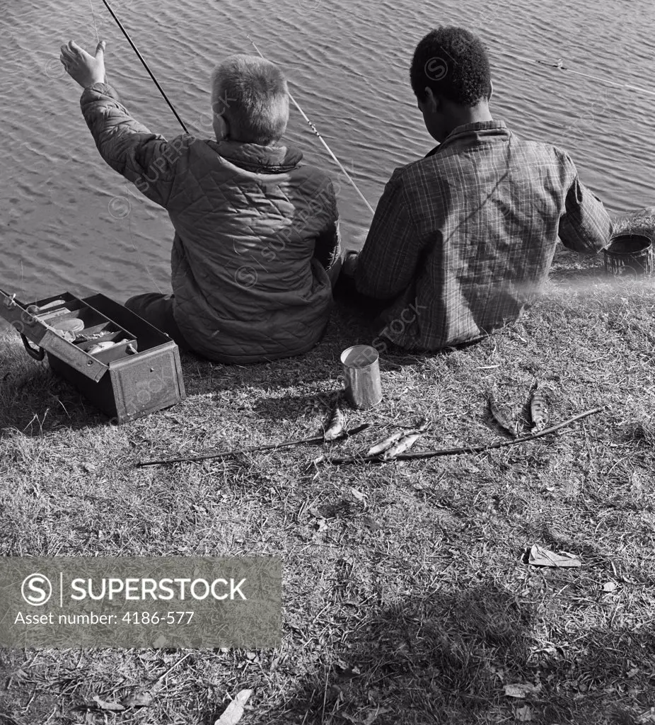 1960S Back View Of Black Boy And White Boy Fishing In Water With Can Of Worms & Tackle Box Shared Together