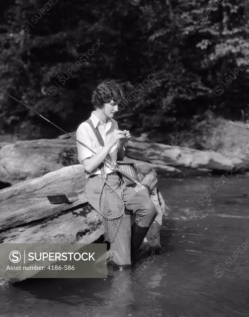1920S 1930S Woman Sitting On Rock By Side Of Stream Wearing Waders Putting Fly On Tippet