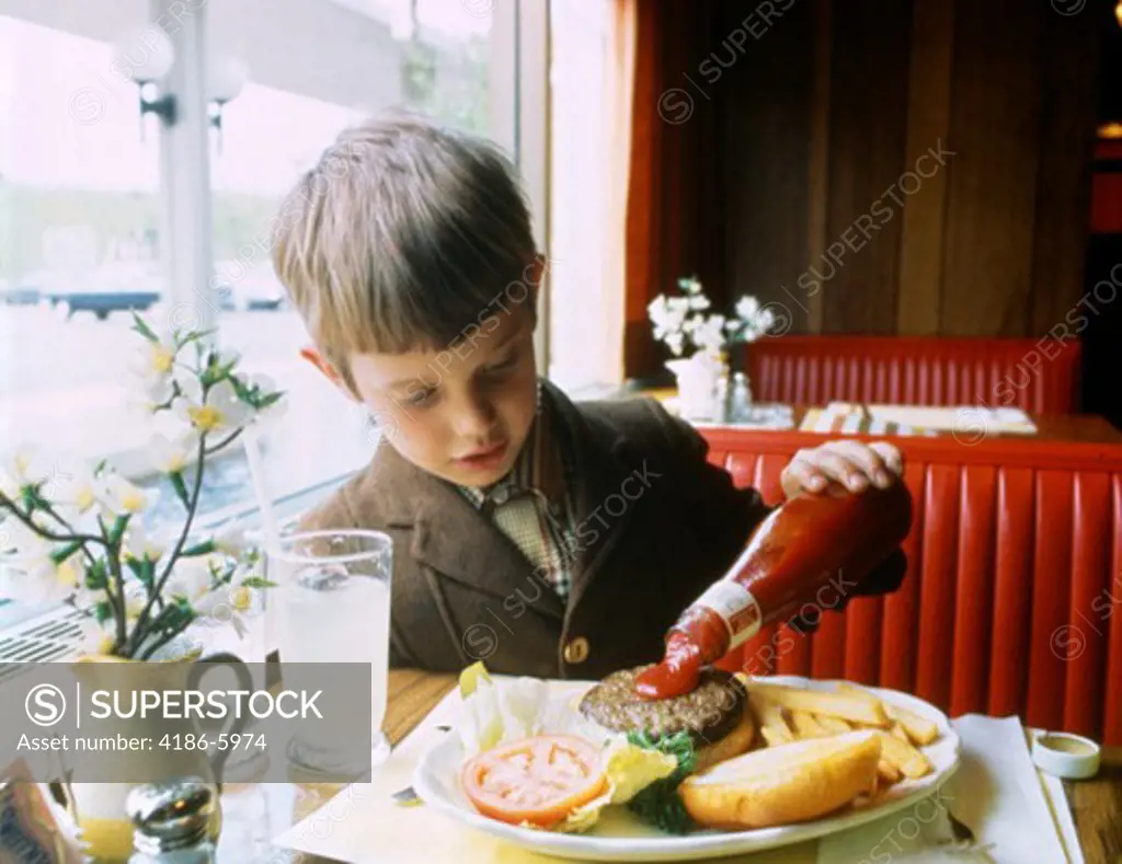 1960S Boy Eating At Booth In Restaurant Pouring Ketchup On Hamburger