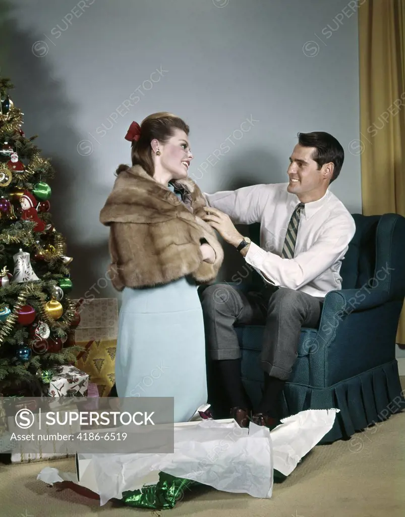 1970S Lifestyle Couple Man Adjusting New Holiday Gift Fur Jacket On Wife In Living Room Beside Christmas Tree