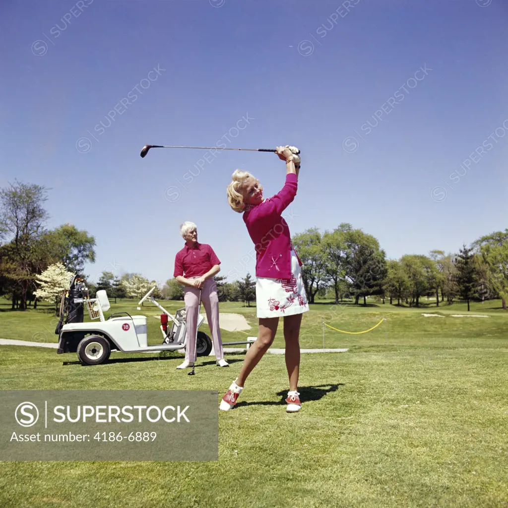 1960S 1970S Mature Woman Swinging Golf Club As Man Stands By Golf Cart