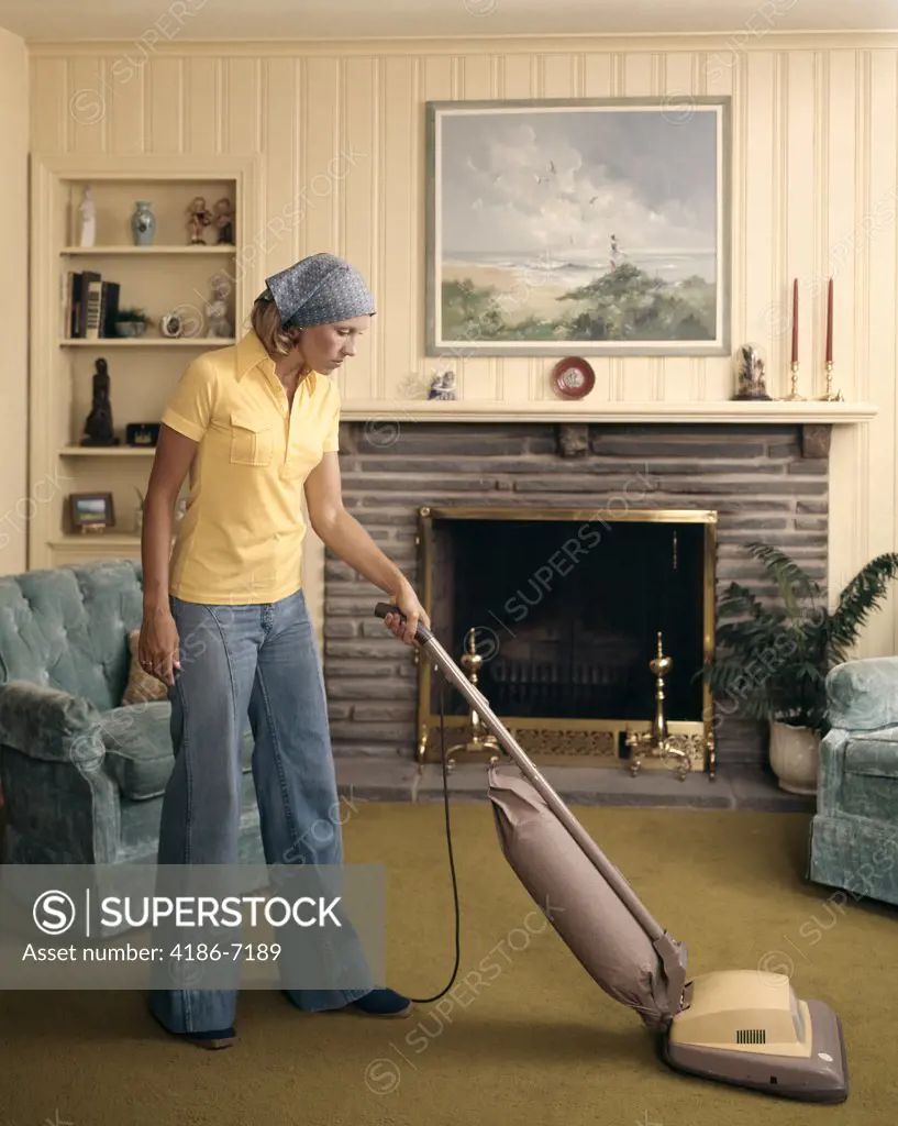 1960S Woman Wearing Head Scarf And Bell Bottom Pants Cleaning Living Room With Upright Vacuum Cleaner