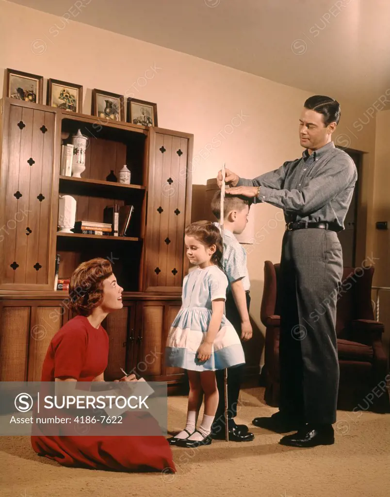 1950S 1960S Family Man Father Woman Mother Boy Son Girl Daughter Measuring And Comparing Height Of Sister And Brother In Living Room
