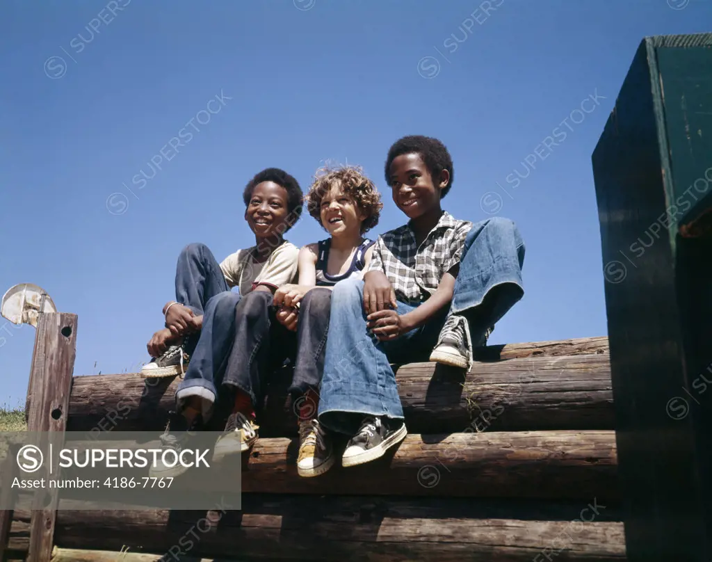 1960S 1970S 3 Boys Sitting Log Fence Smiling Wearing Blue Jeans 2 African American 1 Caucasian  