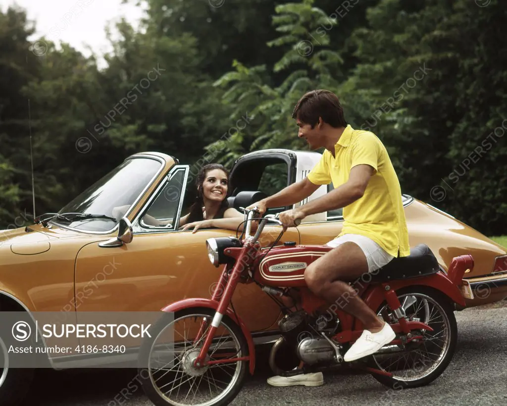 1970S Lifestyle Talking Flirt Couple Man On Small Red Harley Davidson Motorcycle Smiling Woman In Orange Porsche Automobile