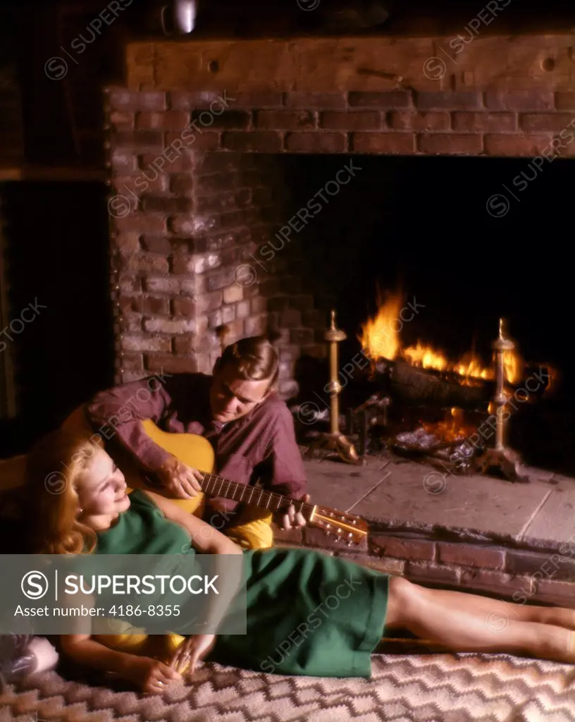 1970S Romantic Couple Man Playing Guitar For Blond Woman Sitting In Front Of Fireplace