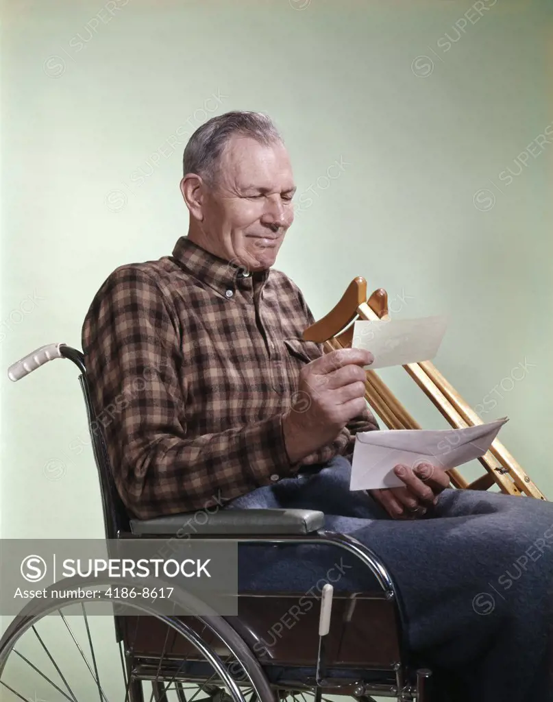 1960S Smiling Senior Man Sitting In Wheelchair With Crutches Reading Insurance Check