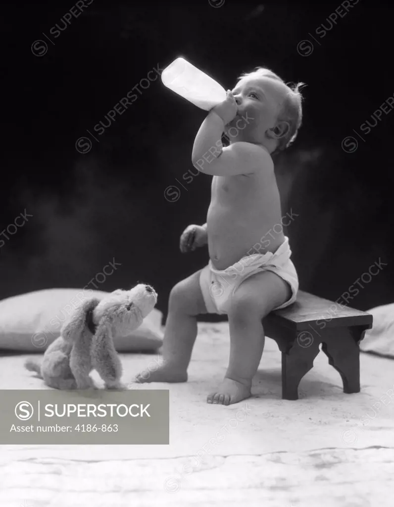 1930S Baby Seated On Stool Drinking Milk From Bottle With Stuffed Puppy Looking Up At Him