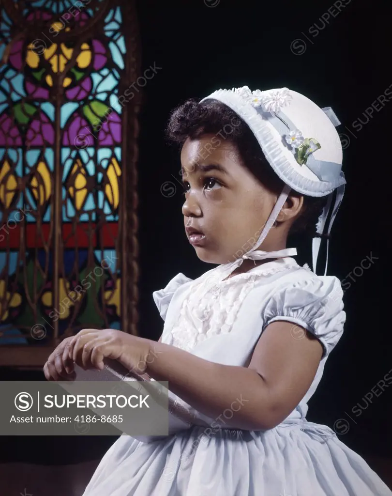1960S Religious Little African American Girl Wearing White Hat And Dress Holding Bible Standing By Stained Glass Window In Church