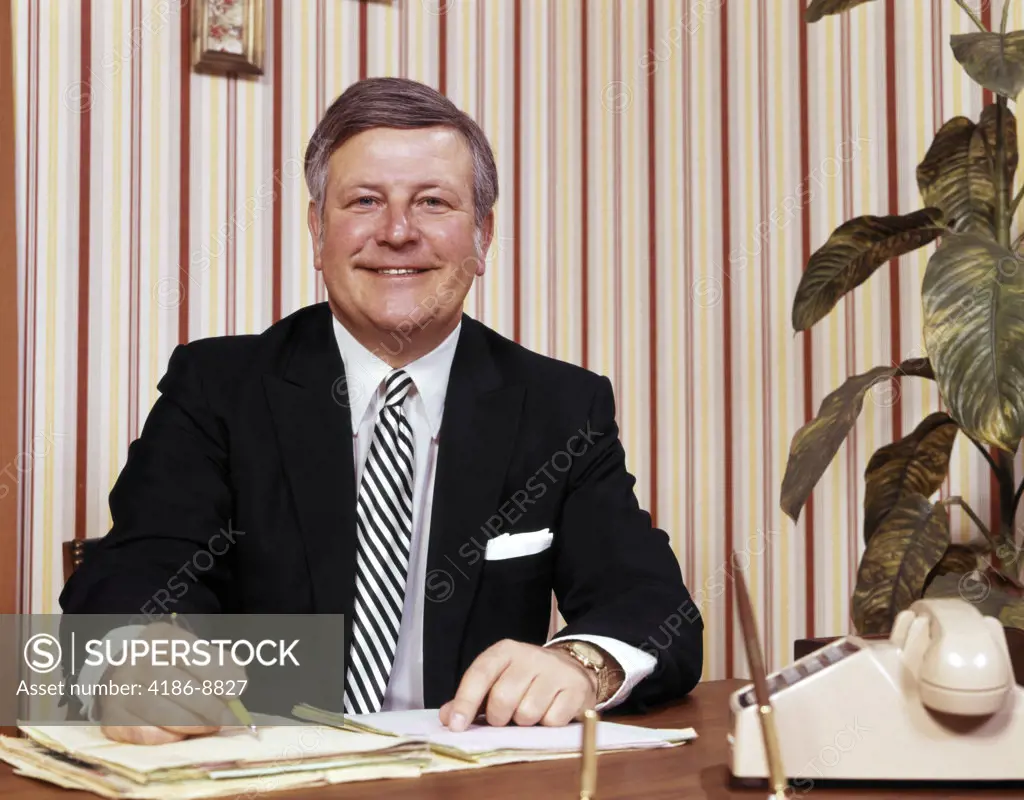 1970S Smiling Businessman Sitting At Desk Writing Papers Folders