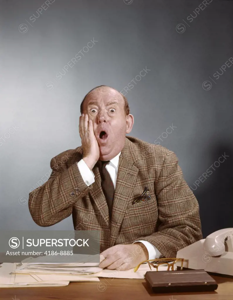 1960S Shocked Balding Executive With Hand To Cheek Sitting At Desk