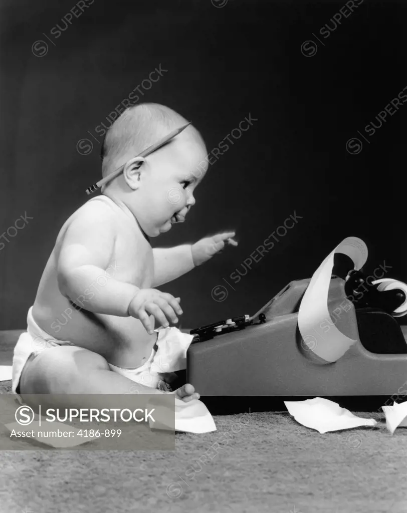 1960S Side View Of Chubby Baby Seated Behind Adding Machine With Pencil Behind Ear & Tape Scattered On Floor